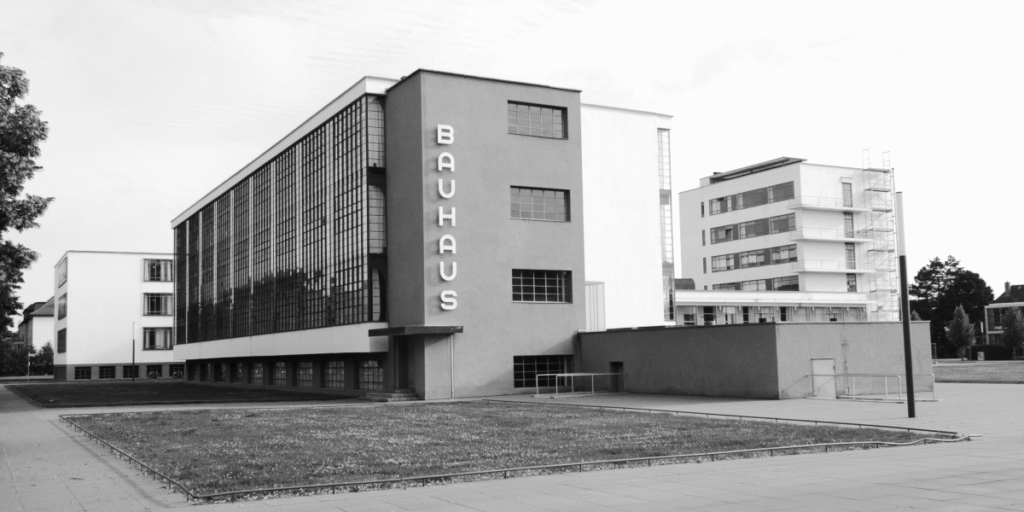 A black and white photograph of the original Bauhaus building in Dessau, Germany. The building is a modernist structure with large windows, clean lines, and geometric shapes.