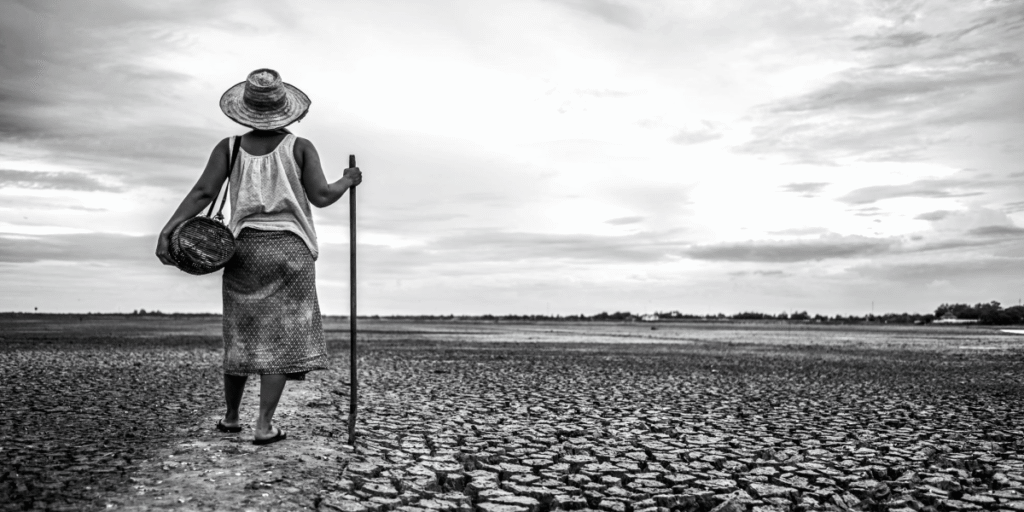 A person wearing a wide-brimmed hat, carrying a basket and a stick, walking across a vast, cracked, dry landscape. A symbolic image of climate change, illustrating why a mobile lifestyle offers resilience.