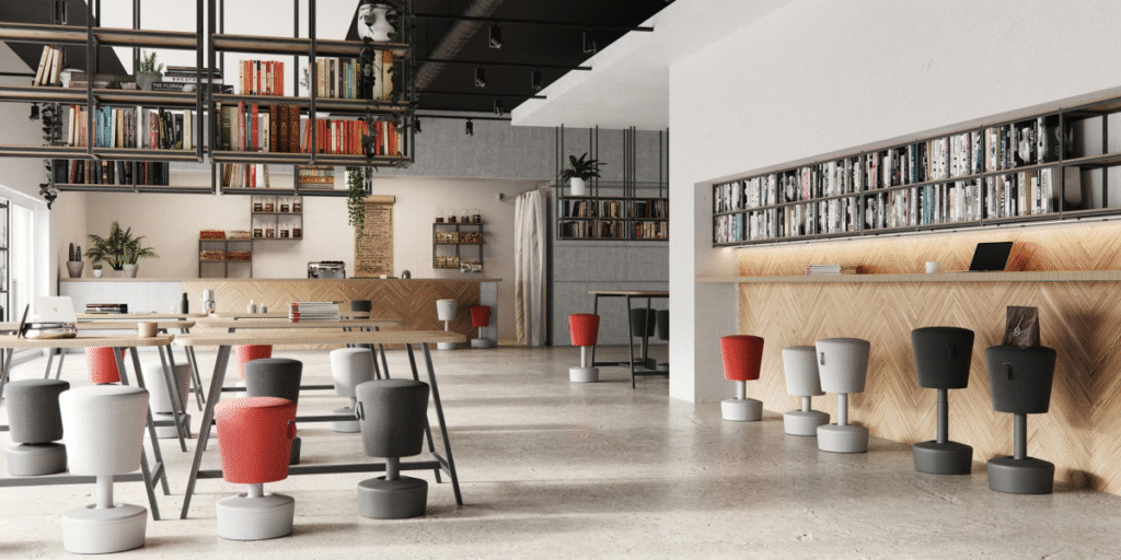 A bright, open communal space with high ceilings, a coffee bar, and multiple tables with colorful, modern-looking stools. Bookshelves line the back wall.