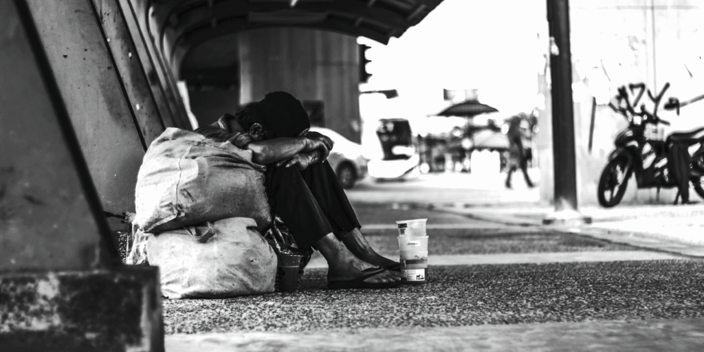 A person sits on the ground in a black-and-white urban setting, hunched over next to a bag, with a cup on the ground.