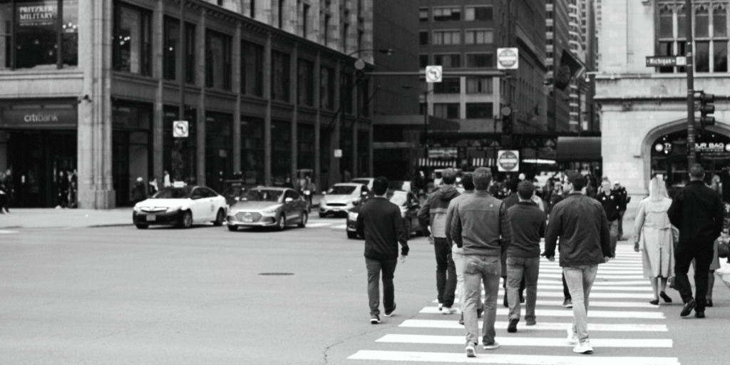 A group of people walking on a city crosswalk in black and white, with cars and buildings in the background. The scene suggests a busy, urban environment.