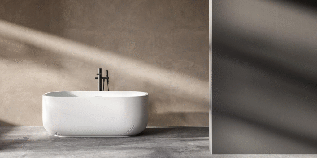 A minimalist bathroom with a white freestanding bathtub, a concrete floor, and a textured, beige wall. Natural light streams in from a window, casting long shadows.