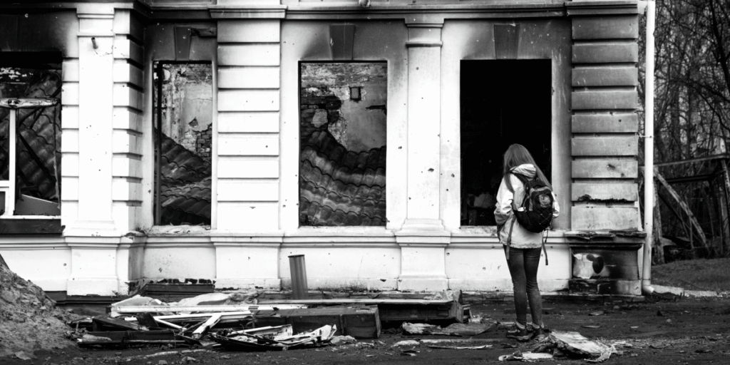 A person with a backpack stands in front of a heavily damaged building, with rubble on the ground, symbolizing the impact of conflict.