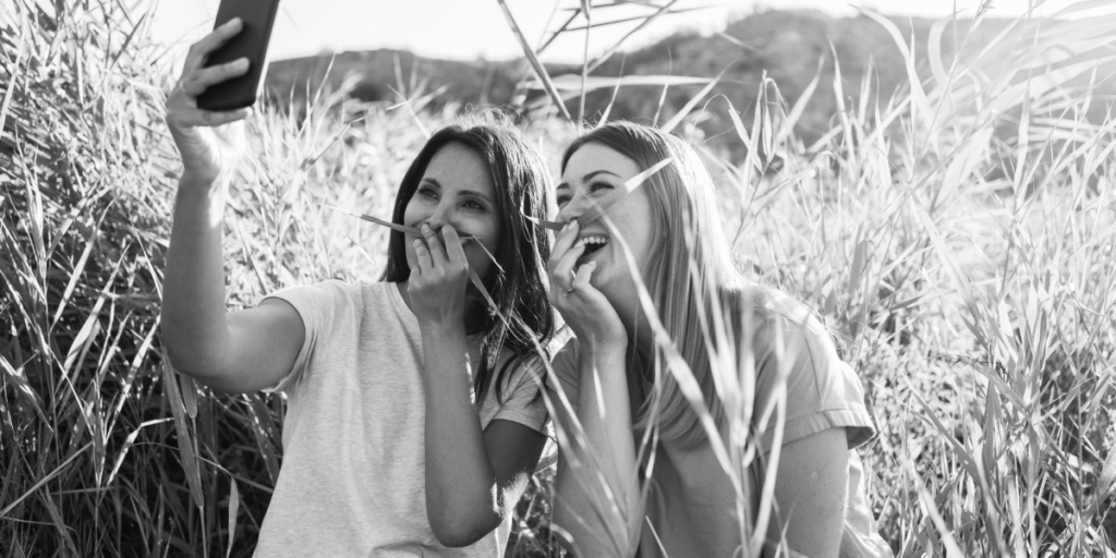 Two women smiling and taking a selfie in tall grass, capturing joy and connection in the present moment.
