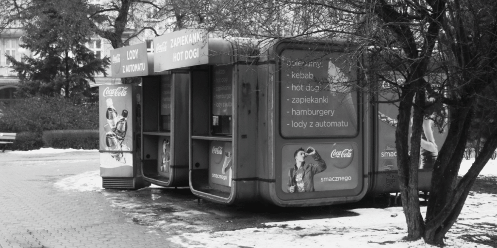 Black and white photo of a K67 kiosk used as a fast food stall with advertisements and menu boards.